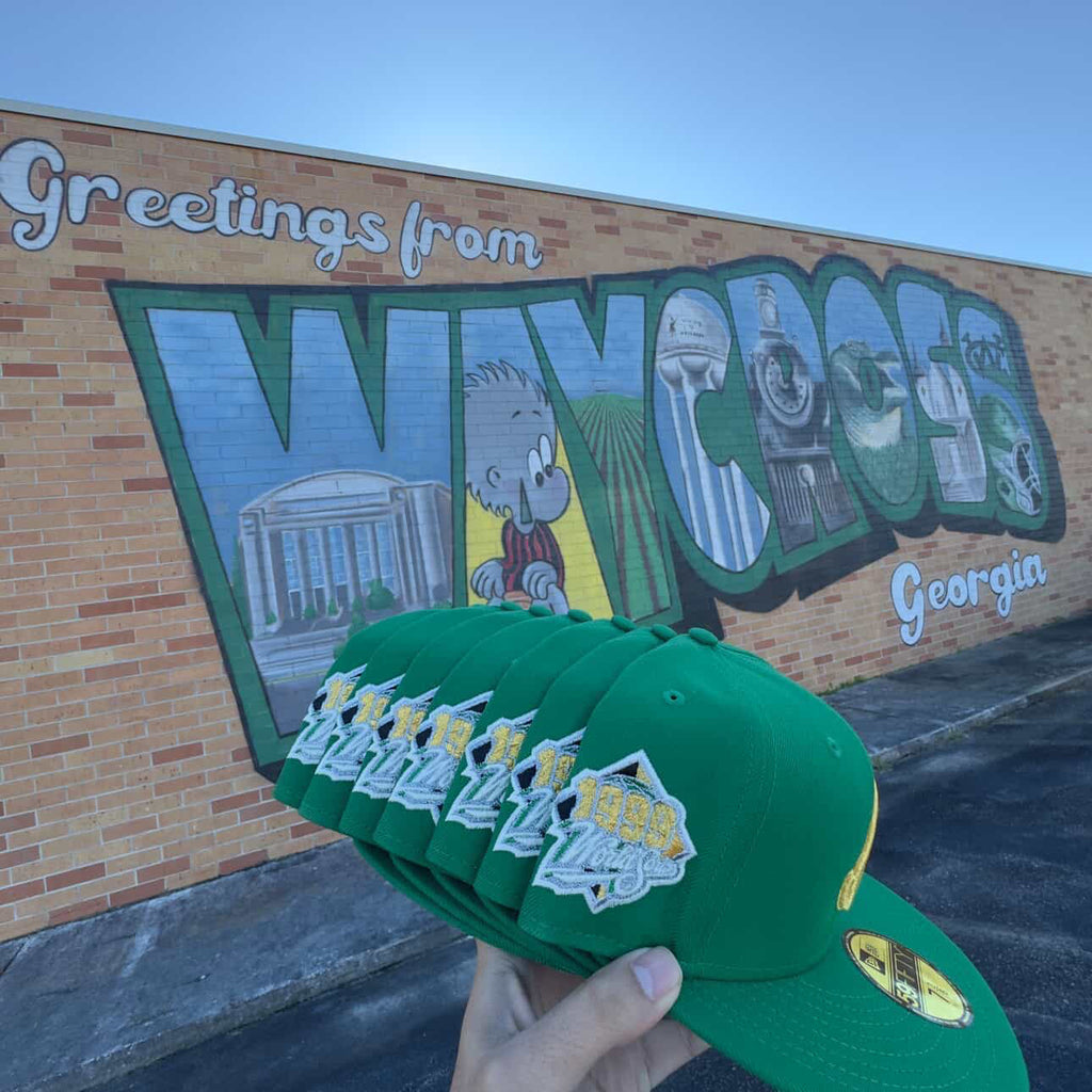 Green New Era fitted caps displayed in front of Waycross Georgia mural, hats, sneakers, footwear