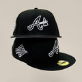 A black Atlanta Braves fitted cap with white embroidered logos, shown from multiple angles against a solid beige background.