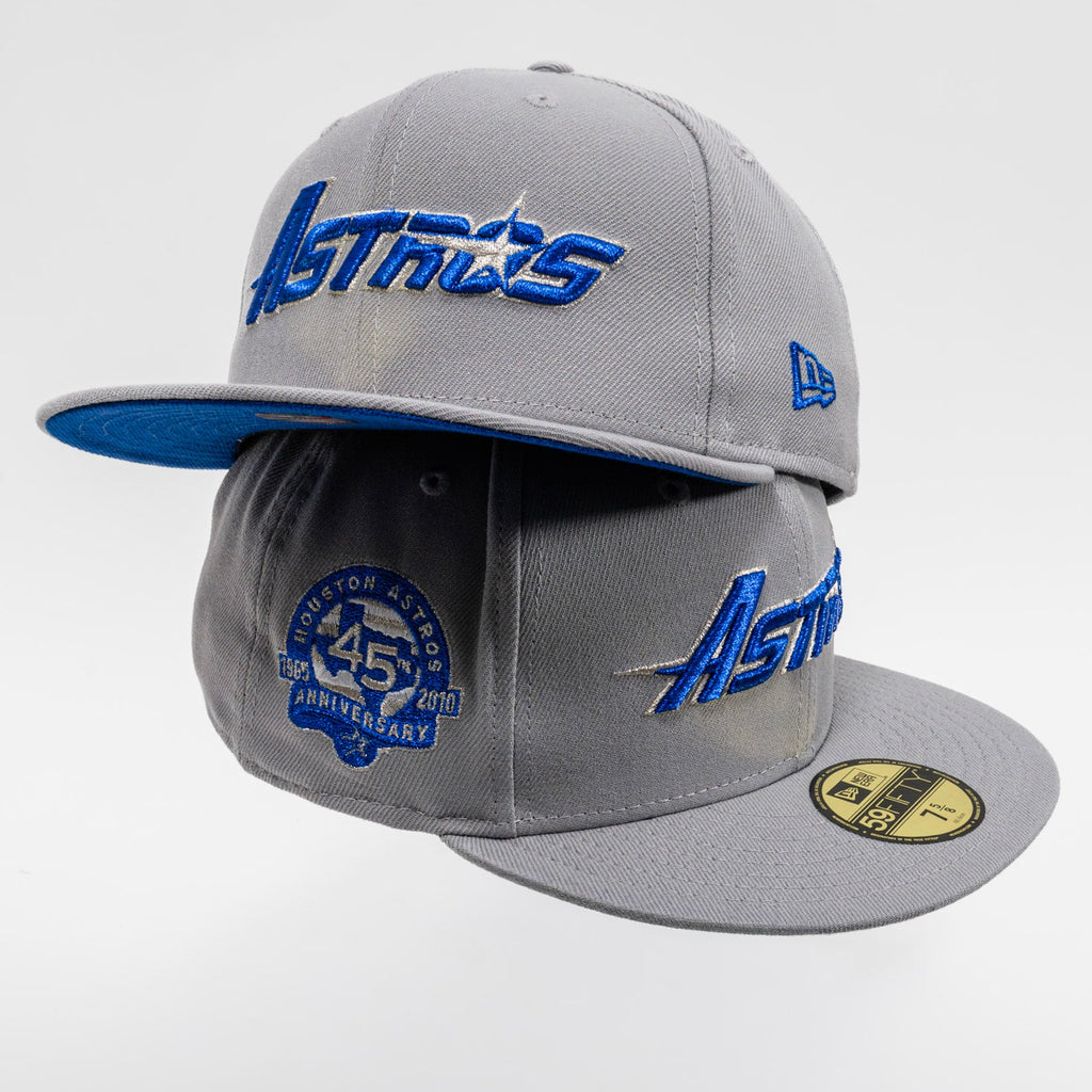 Two gray Houston Astros baseball caps stacked together on a light gray background. The top cap features blue embroidered 'Astros' lettering on the front and a blue side logo, while the bottom cap shows a blue 45th Anniversary patch on the front and a gold New Era 59FIFTY sticker on the brim.