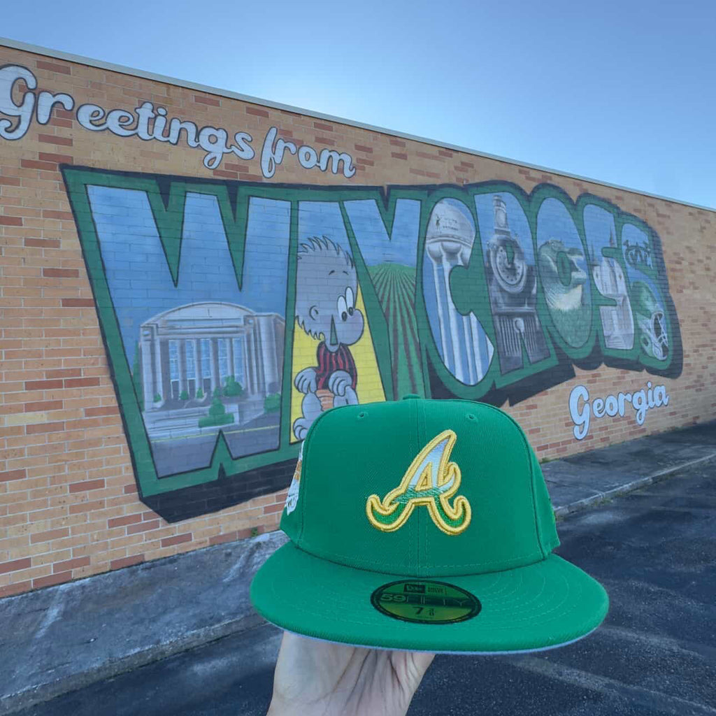 Green New Era fitted cap with yellow A logo held in front of Waycross Georgia mural, hats, fitted caps